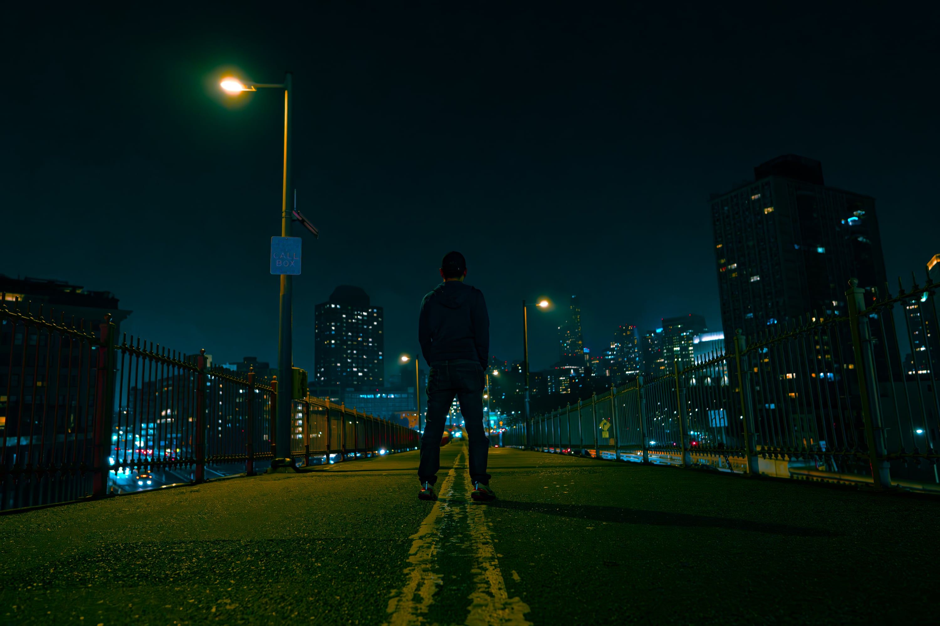 A lone figure standing on a bridge walkway at night, facing the city skyline